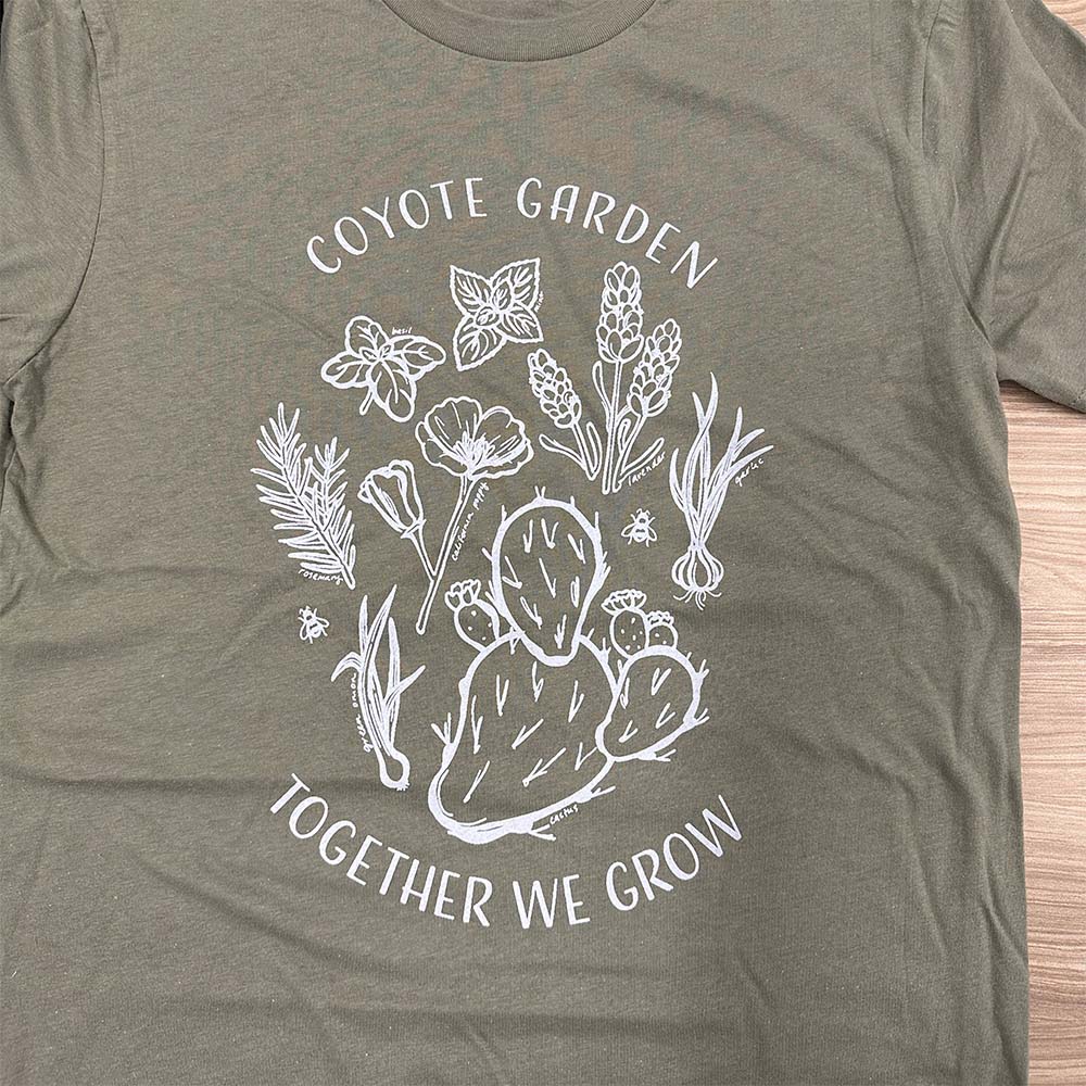 White line drawing on a green T-shirt. The drawing shows an arrangement of basil, mint, lavender, rosemary, a California poppy, garlic, green onion, and cactus. Text on the shirt reads: Coyote Garden Together we grow.